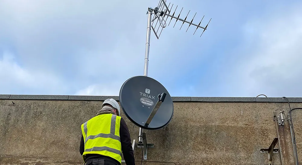 Installer in high-visibility clothing fitting a satellite dish and TV aerial to a wall-mounted pole